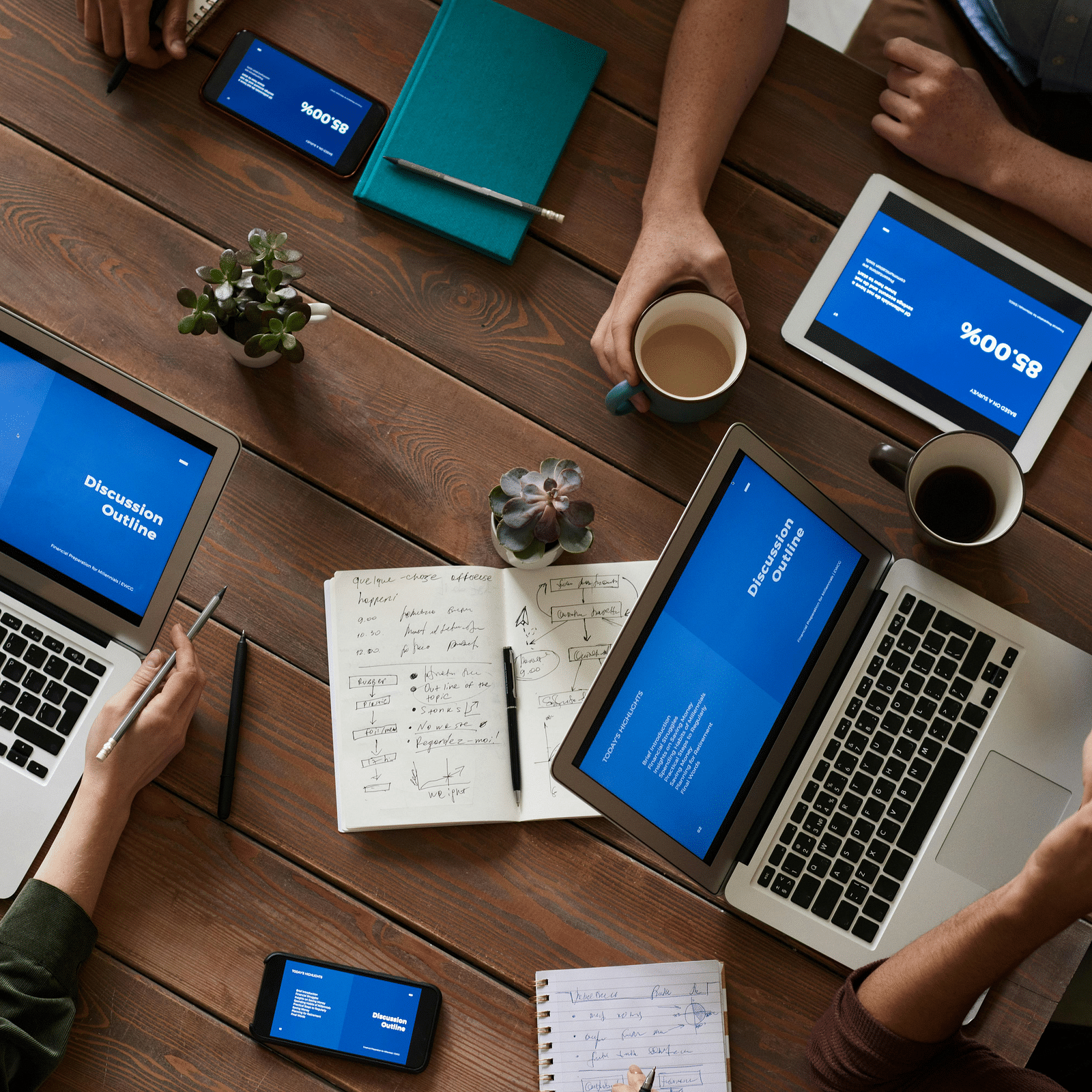 A top-down view of a table featuring people working on laptops, surrounded by notes, coffee mugs, and phones nearby