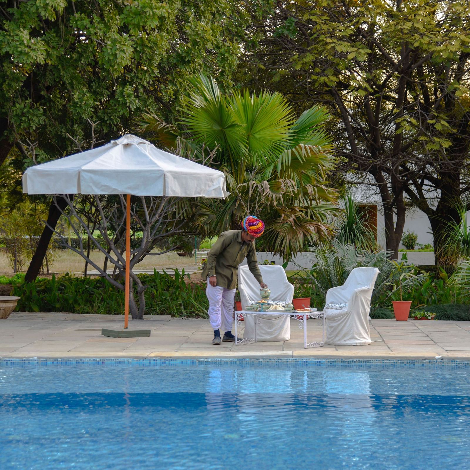A person in a turban arranges items on a table beside a swimming pool, with a parasol and seating nearby, amidst lush greenery- Shahpura Bagh.