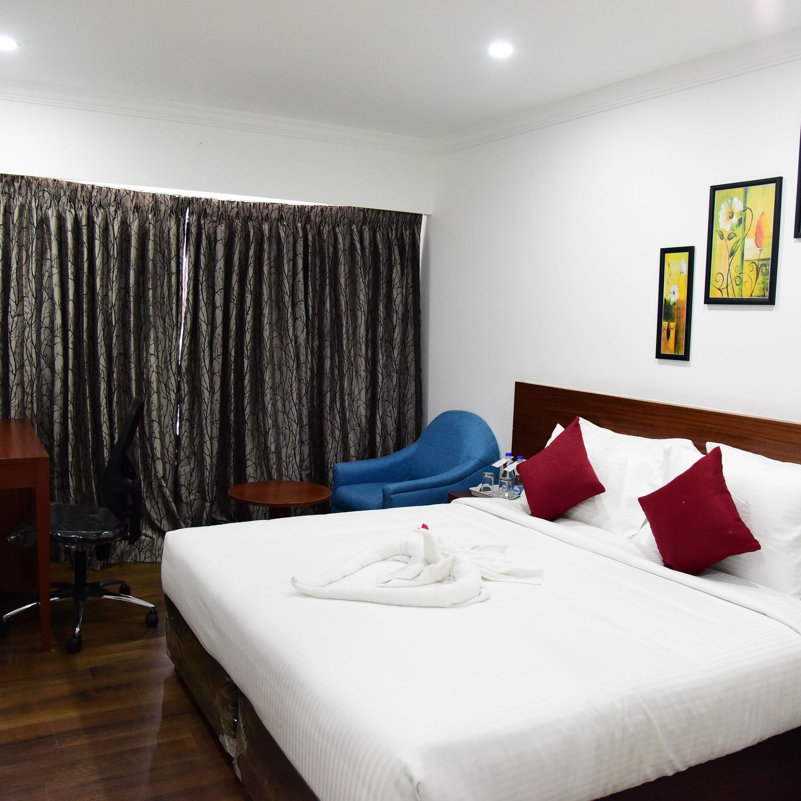  A spacious royal deluxe room at The Royal Comfort, featuring a king-sized bed with rich wooden accents, a deep red runner, and a modern painting above the headboard.