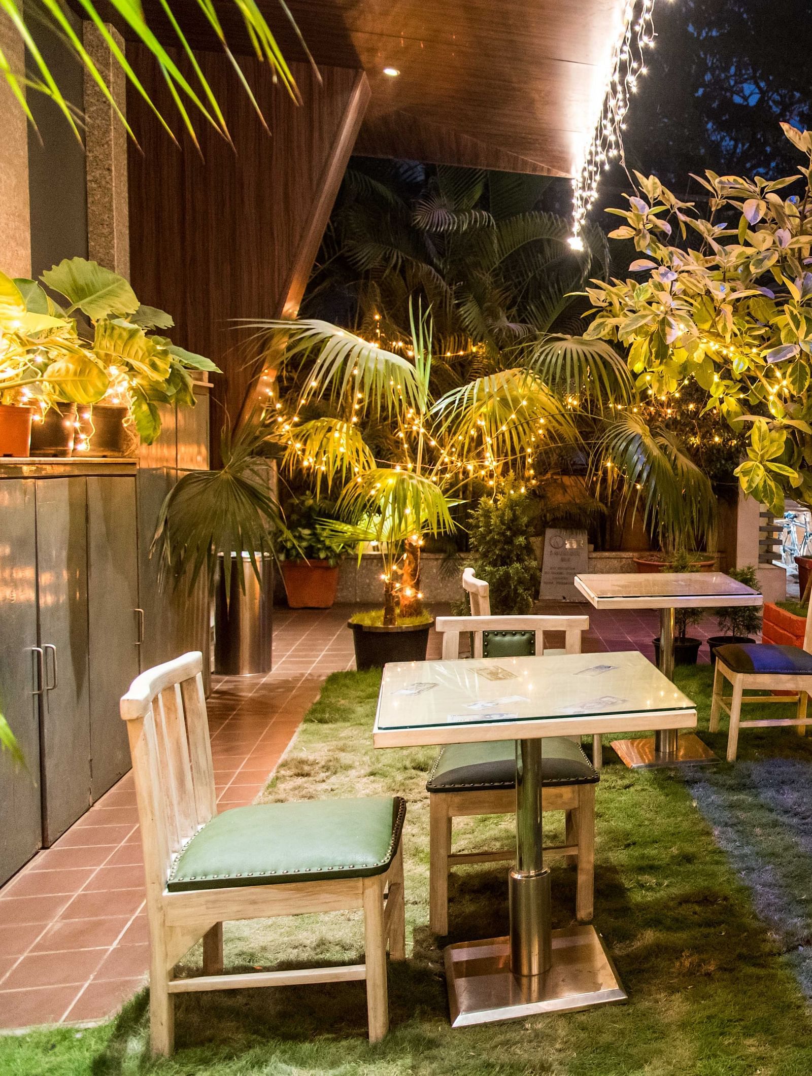 Cosy outdoor dining with wooden tables, chairs, and lush plants illuminated by warm lighting.