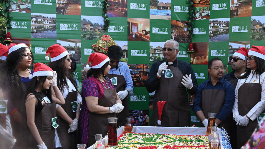 A group of staff and guests wearing Santa hats gathered around a large decorated Christmas cake at Vedic Village Spa Resort during a festive Christmas celebration event.