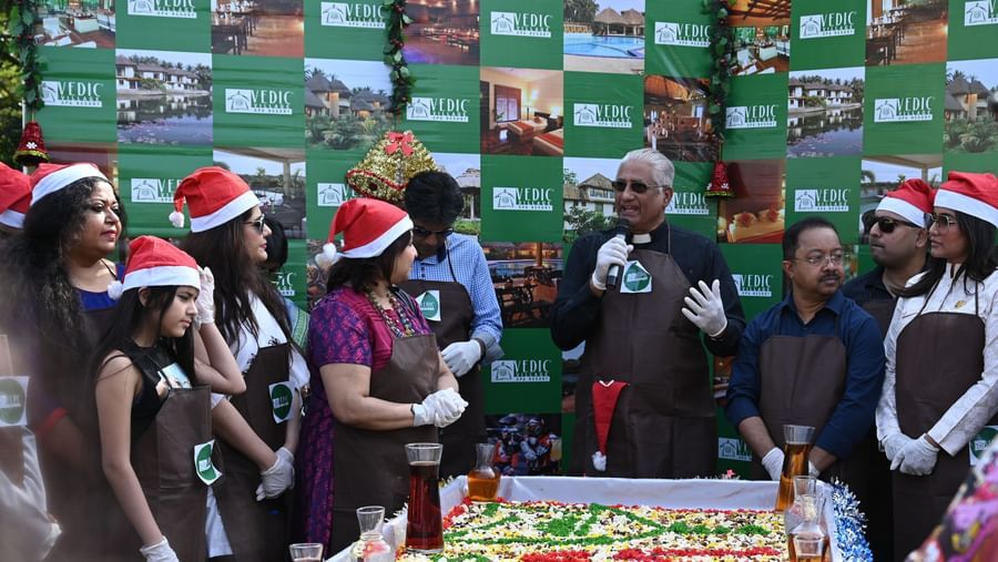 A group of people wearing Santa hats gathered around a table with a large cake or food display | Vedic Village Spa Resort, Kolkata
