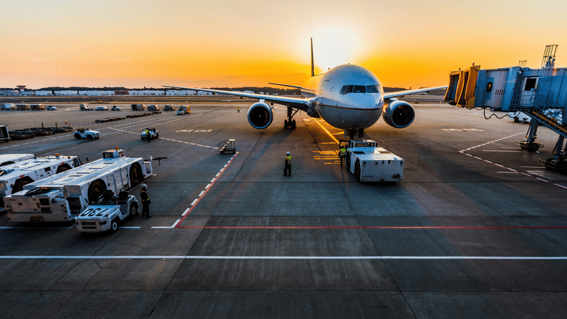 An aeroplane on the runway of an airport surrounded by other vehicles against a orange sky with the sun setting.