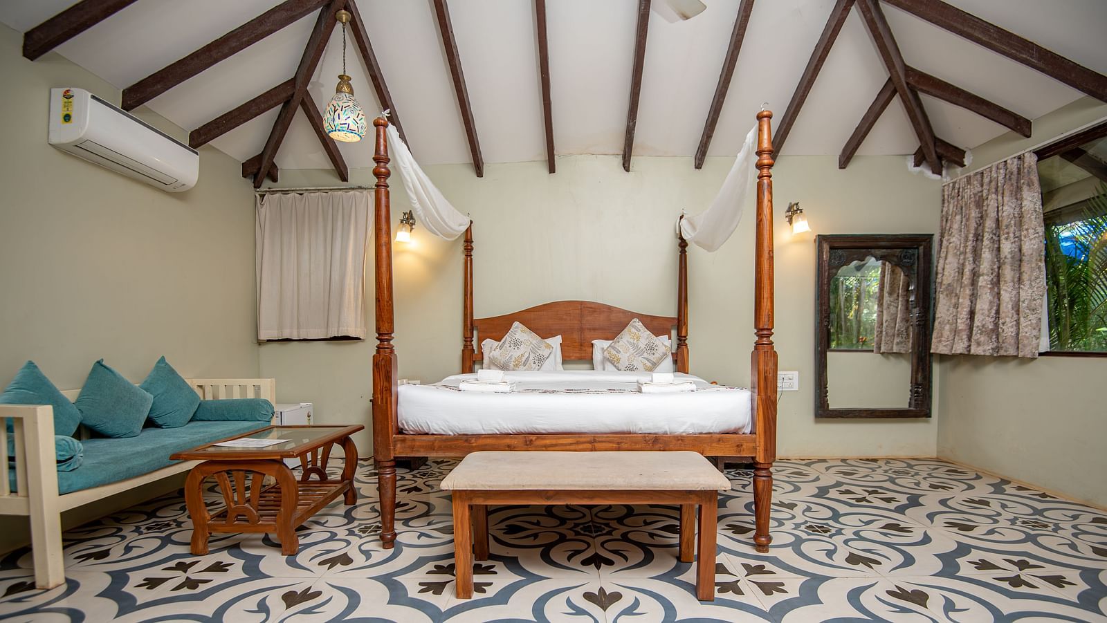 Spacious room with a large four-poster bed, cushioned sofa and wooden table, and a side window with curtains pulled aside at De Soul Santé, Morjim.