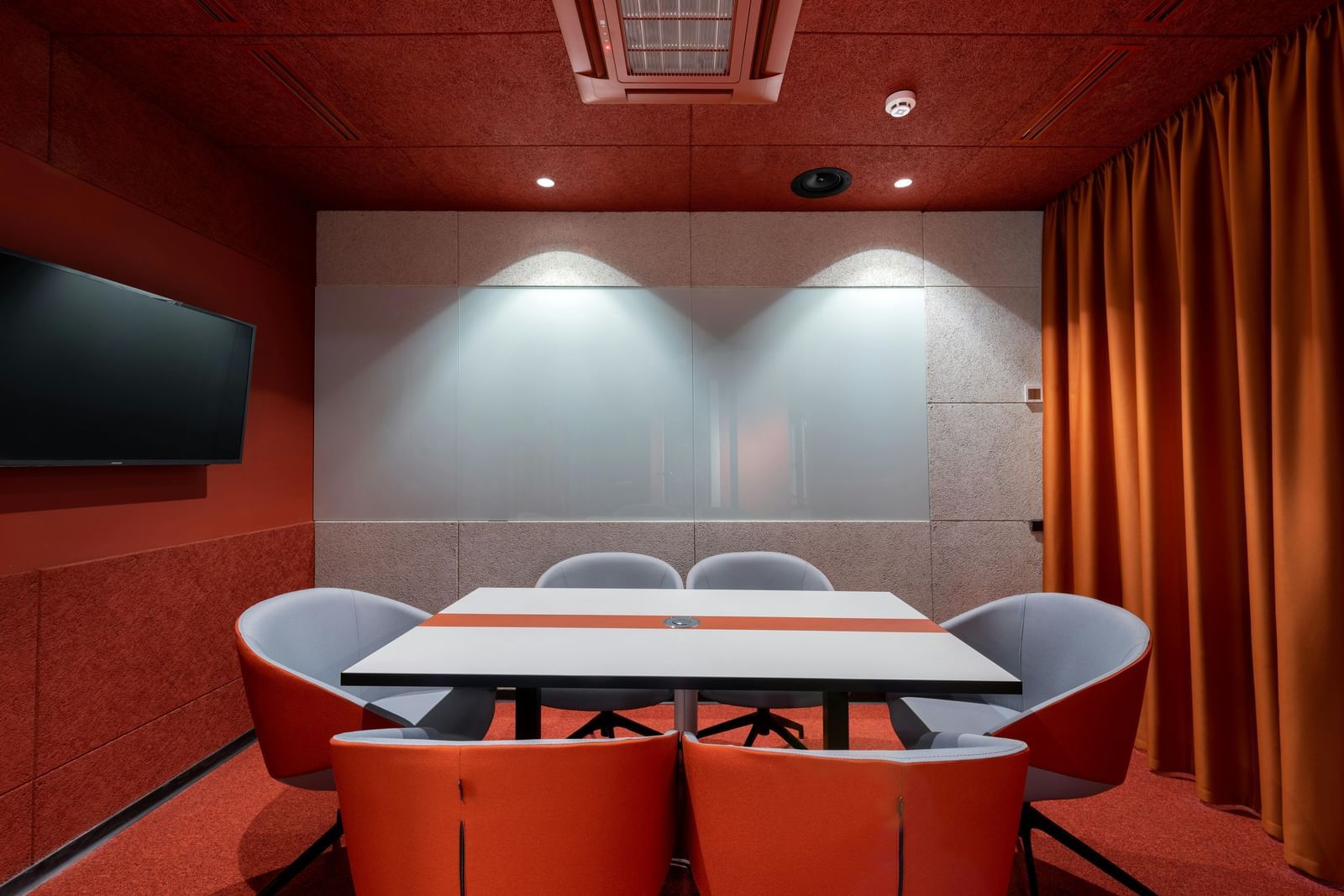 A modern conference room with red walls, a white table surrounded by grey and orange chairs, and a large screen on the left wall.