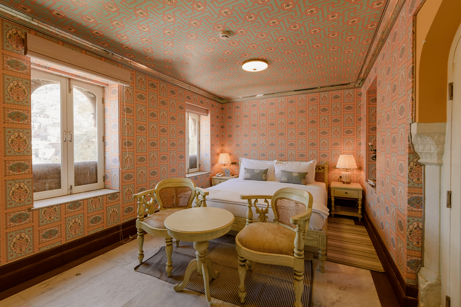 Deluxe rooms in Surya Haveli Jaipur featuring a sitting space in front of the plush bed with coffee table and a couple of chairs near the fancy window