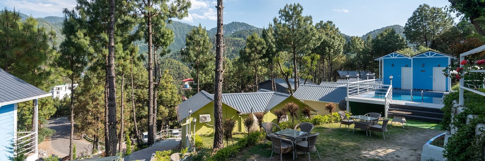 Scenic hillside resort with pine trees, outdoor seating area, and blue-roofed cottages set against a backdrop of lush green mountains under a clear sky - DLS Nature Trinket Resort, Dalhousie.
