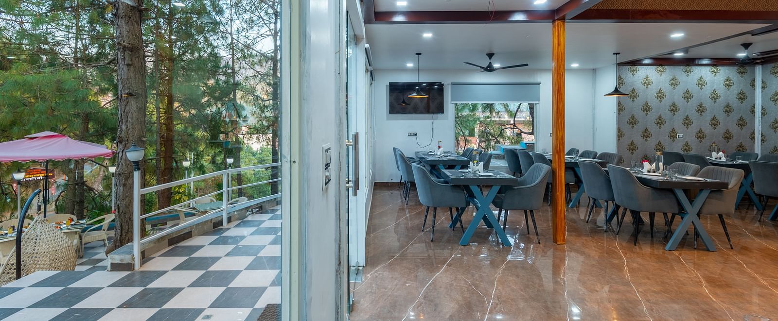 The interior of a restaurant is shown with tables and chairs. Through the open doorway, there's a view of a hillside - DLS Nature Trinket Resort, Dalhousie.
