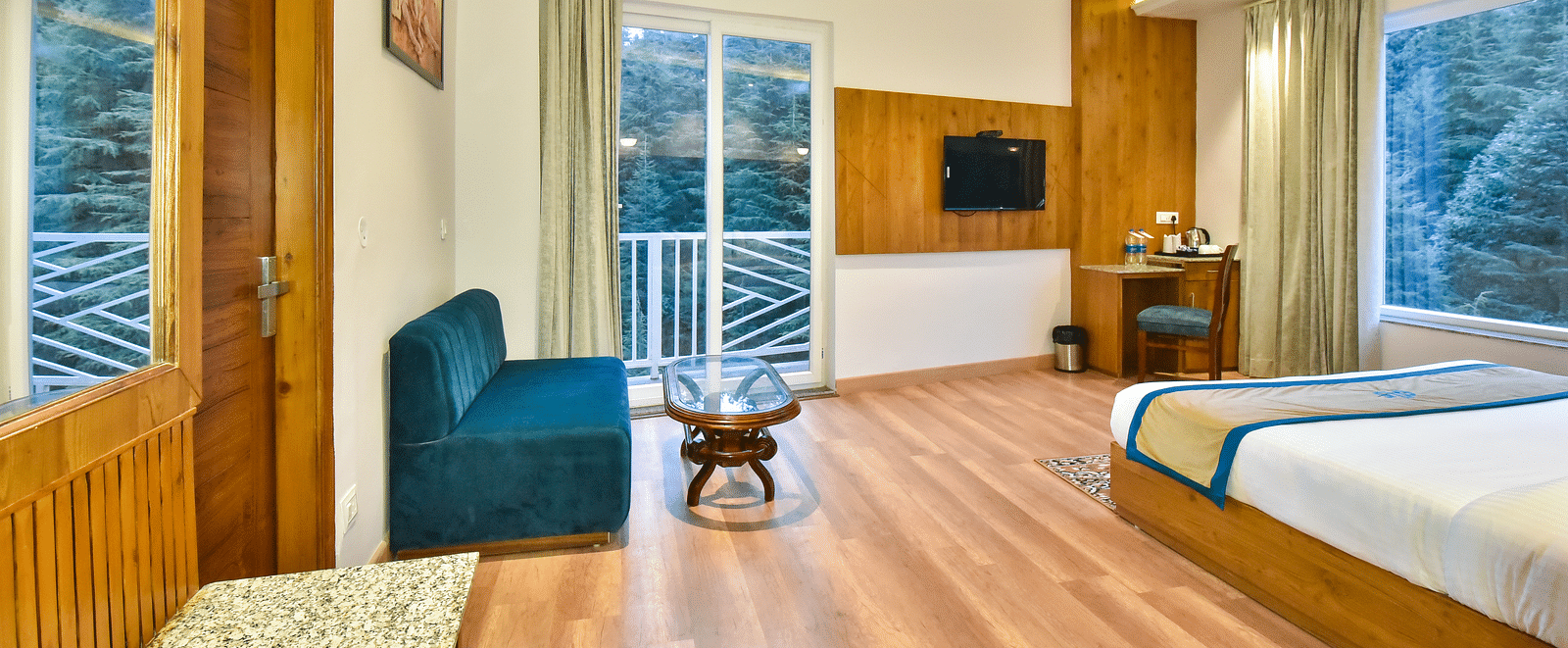 A spacious luxury room with balcony featuring a bed, a couch, a coffee table, flat screen TV, a wall mounted mirror, and an expansive glass door leading to the balcony - DLS Ark Holiday Inn, Dalhousie