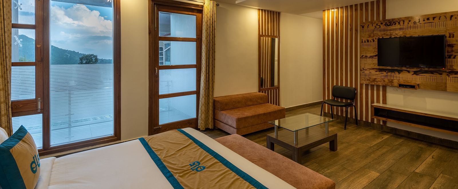 A premium room featuring a comfortable bed, wooden decor, flat-screen TV, and windows offering a scenic view for a relaxing stay - DLS Meghavan Resort, Dharamshala