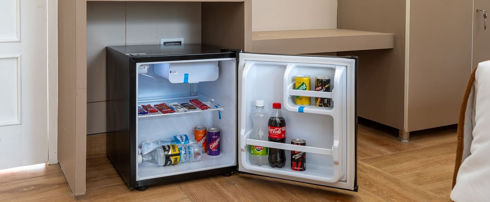  Small open minibar fridge in a hotel room, stocked with beverages and snacks, with a red electric kettle, cups, and complimentary items on the counter above - DLS Nature Trinket Resort, Dalhousie.
