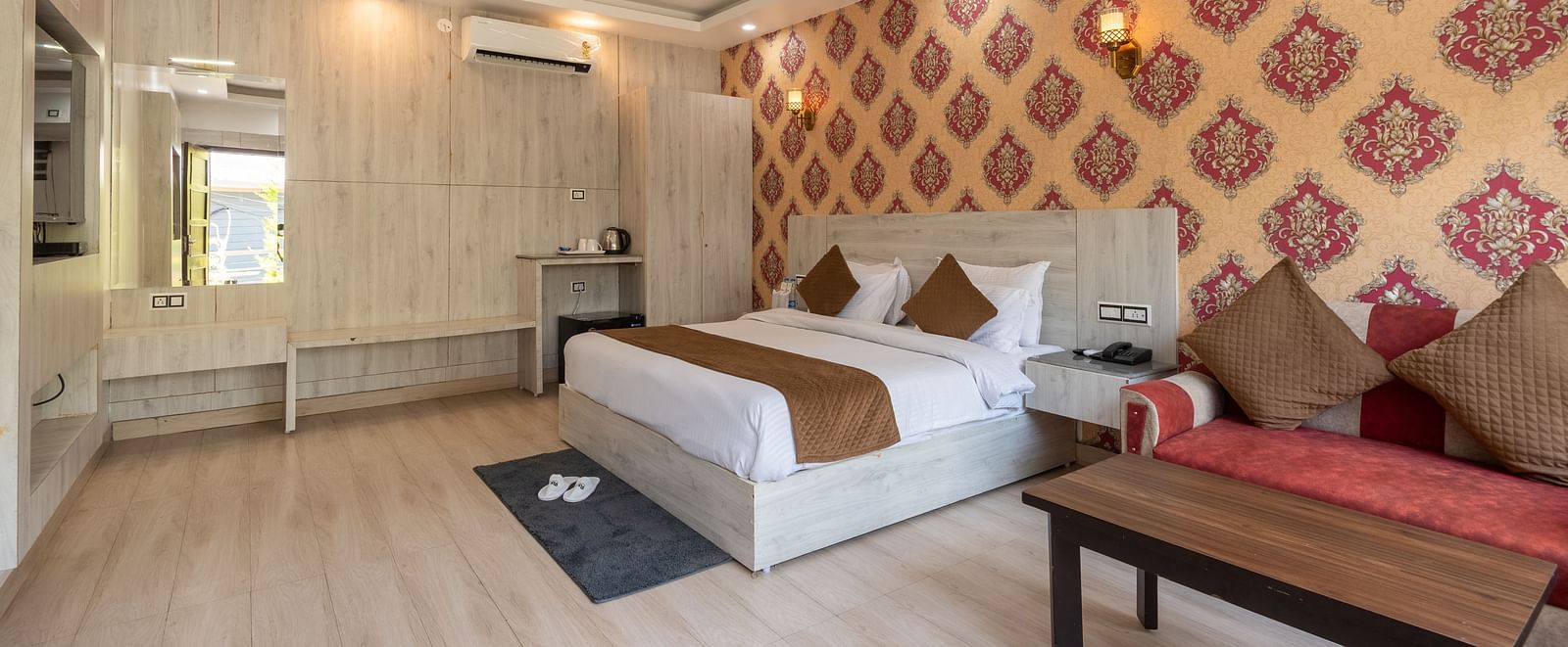Spacious, well-lit hotel room with a large double bed, patterned feature wall in red and gold, modern furnishings, ceiling fan, and a comfortable seating area with a coffee table - DLS Nature Trinket Resort, Dalhousie.