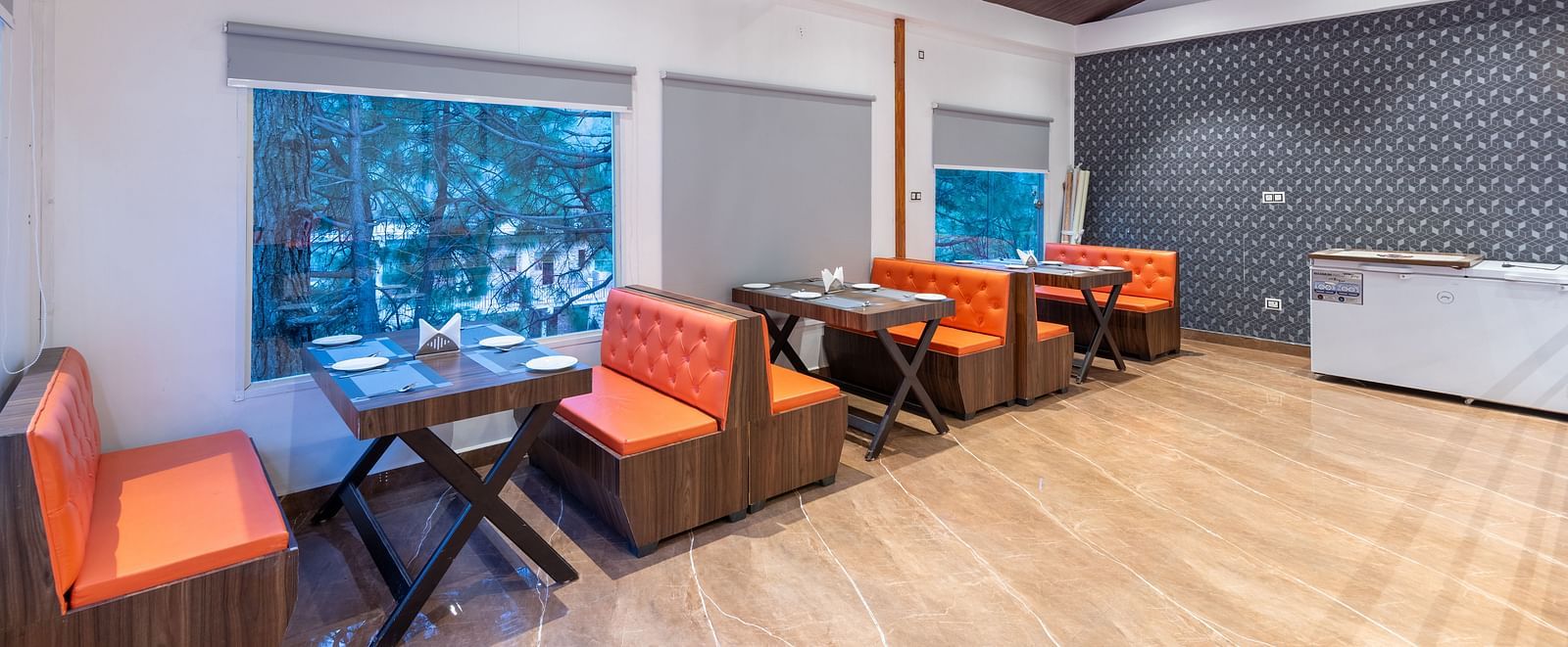 A brightly lit restaurant interior features wooden floors, a dark brown ceiling, tables with place settings, orange cushioned booths and chairs, and large windows with grey blinds - DLS Nature Trinket Resort, Dalhousie.