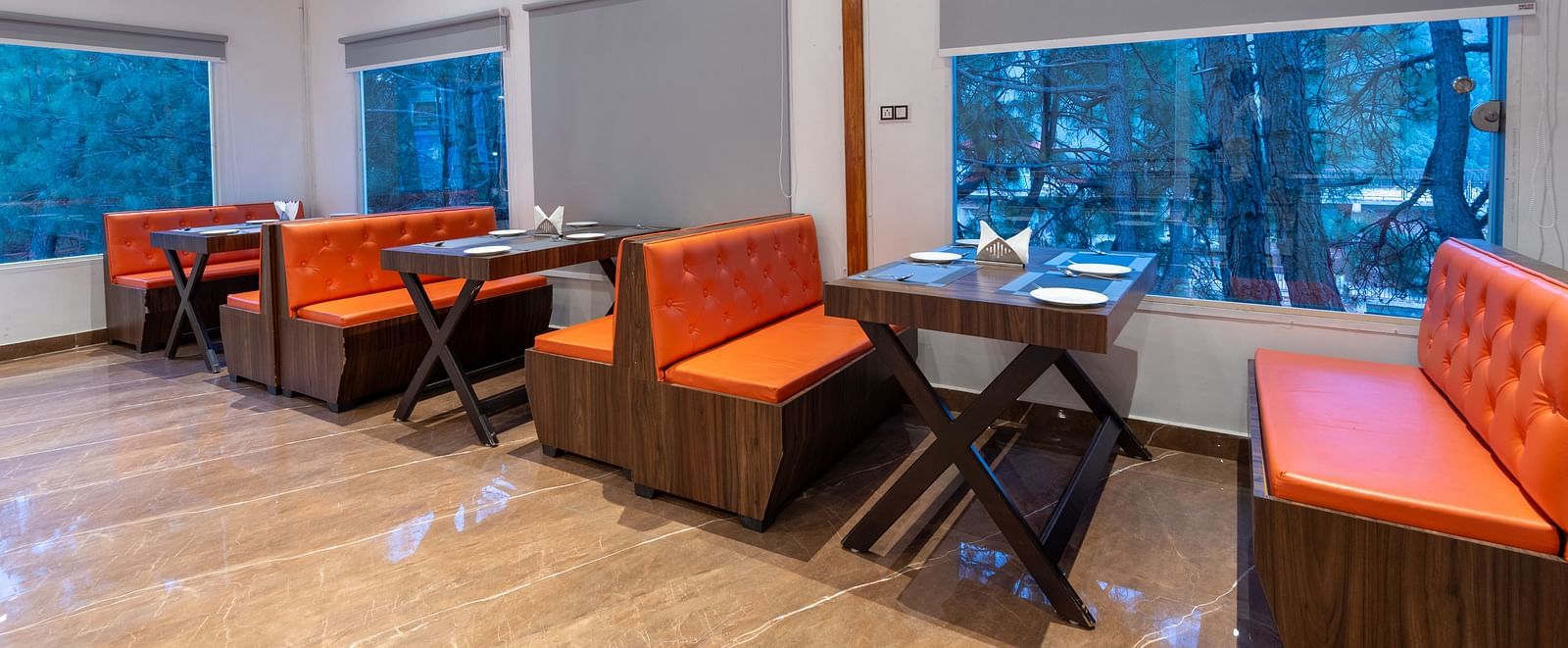 A brightly lit restaurant interior features wooden floors, a dark brown ceiling, tables with place settings, and orange cushioned booths and chairs situated near large windows with grey blinds - DLS Nature Trinket Resort, Dalhousie.