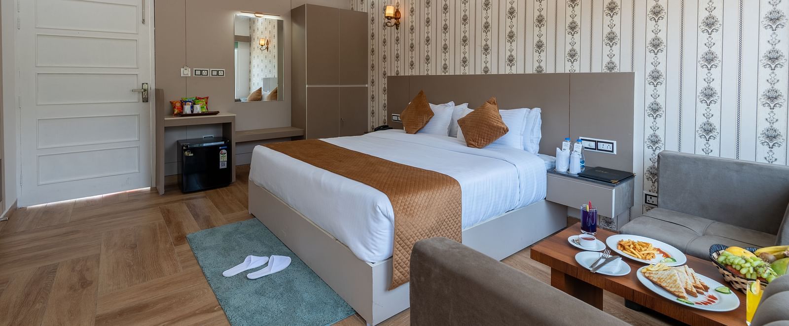 A hotel room with a large bed, modern decor, a seating area with food on the table, and amenities including a ceiling fan, wardrobe, and minibar - DLS Nature Trinket Resort, Dalhousie.