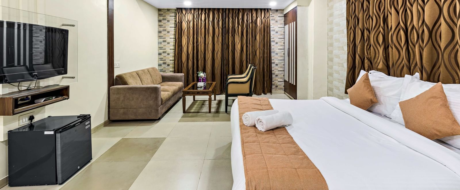 A well-lit suite in Krushnai Resort featuring a double bed with blue runner, a wall mounted TV, docking station, couch and a lounge chair.