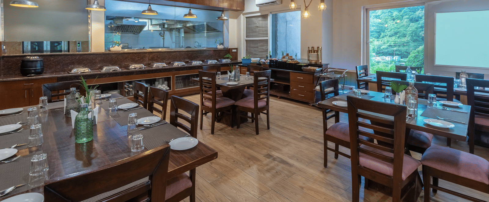 A view of a restaurant at DLS Hotels on the Ganges, Rishikesh with wooden tables, chairs, ceiling lights, and a serving counter in the background.