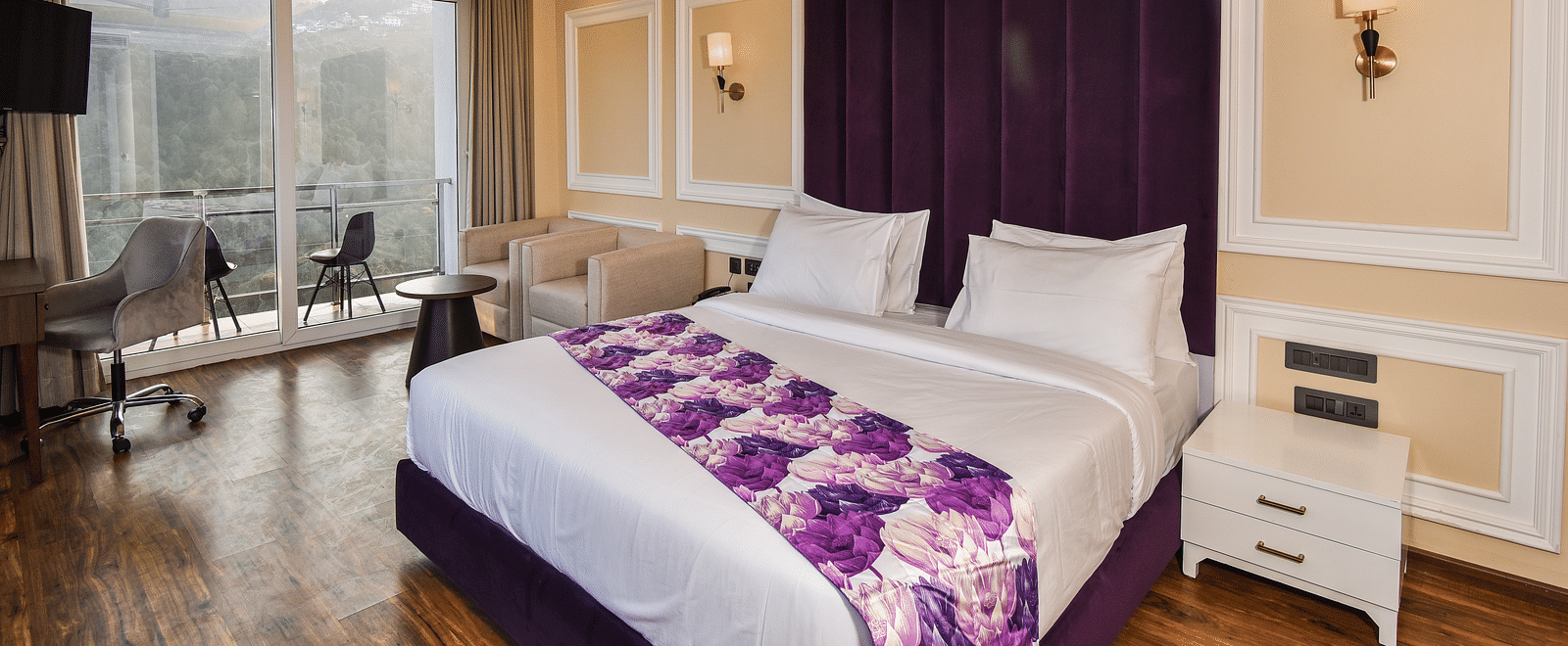 A vibrant bedroom with a large bed featuring purple accents and a bright atmosphere. The room includes large windows that provide scenic views of the hills outside.