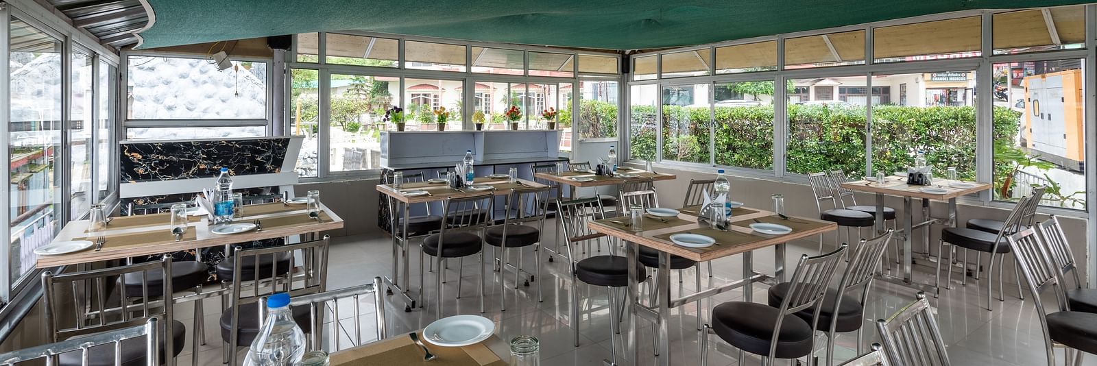 A cafe in Bhagsu with tables and chairs, featuring a green canopy overhead and large windows with views of the outside. The tables are set with plates, glasses, and bottles of water - DLS Meghavan Resort, Dharamshala