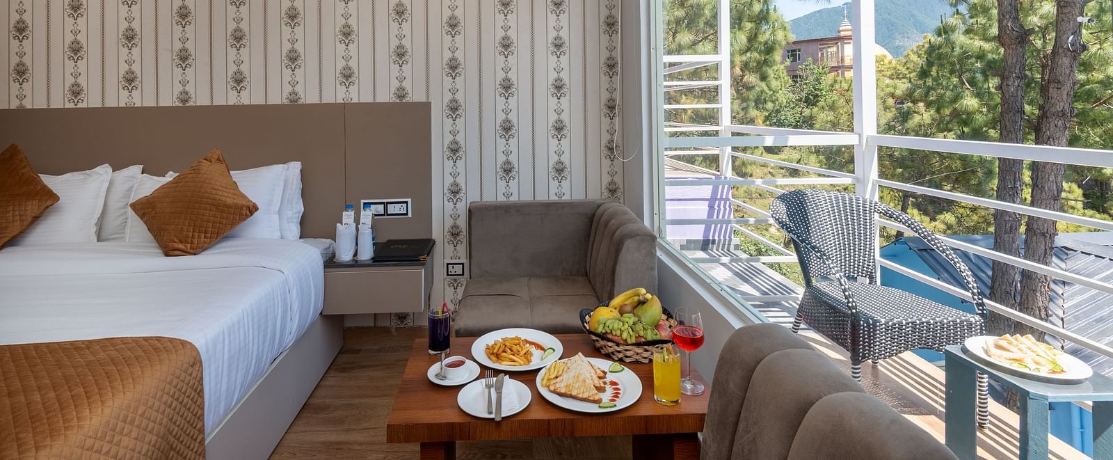 Comfortable hotel room featuring a neatly made bed, a sitting area with food and drinks on a coffee table, and a large window with a view of pine trees and distant mountains from the balcony - DLS Nature Trinket Resort, Dalhousie.