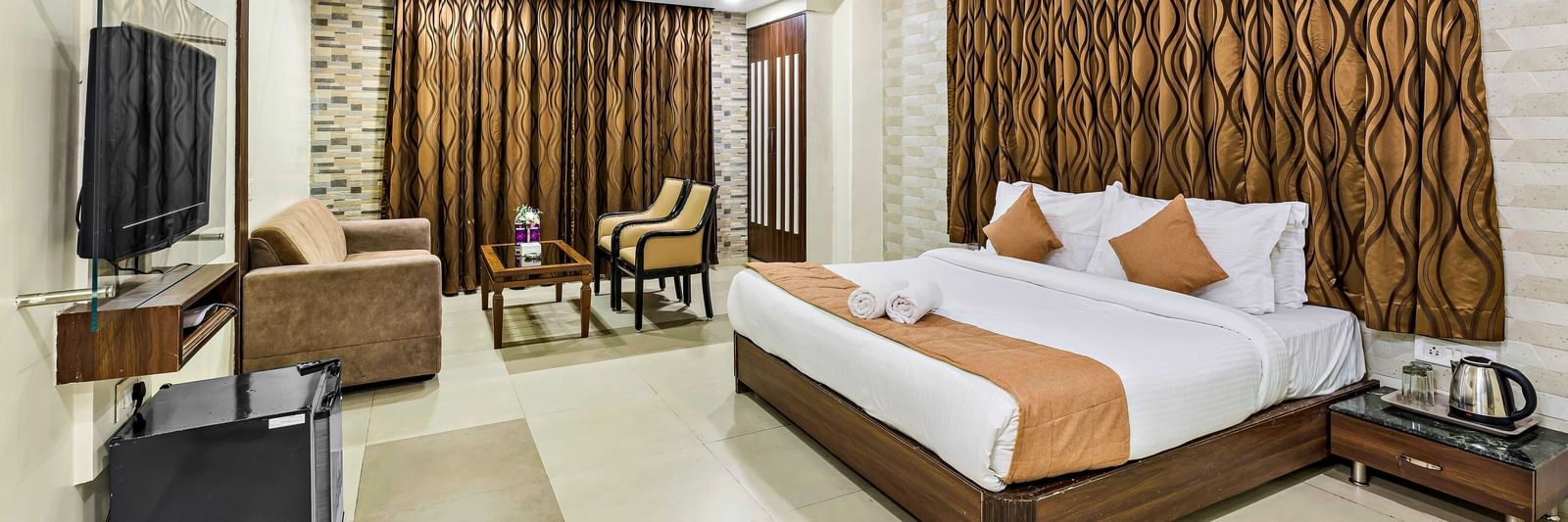 A wide view of a suite in Krushnai Resort featuring a double bed with blue runner, a wall mounted TV, docking station, couch and a lounge chair.