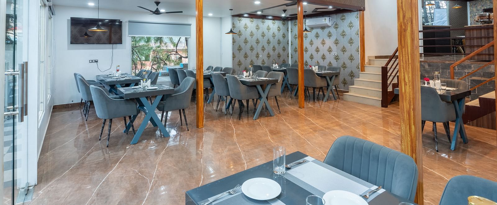 Elegant restaurant interior with grey upholstered seating, set dining tables, tiled flooring, and decorative wallpaper near a staircase - DLS Nature Trinket Resort, Dalhousie.