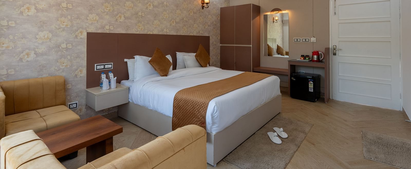 Modern hotel room with a double bed, warm lighting, ceiling fan, cushioned seating area, dressing table, and neatly arranged amenities on the side counter - DLS Nature Trinket Resort, Dalhousie.