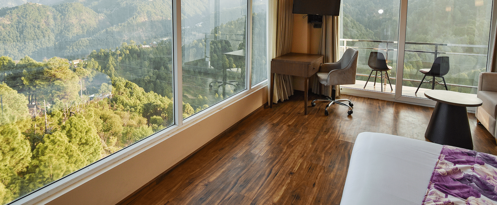 A modern bedroom with a king-sized bed, a seating area by the window, and large glass windows providing expansive views of the mountains.