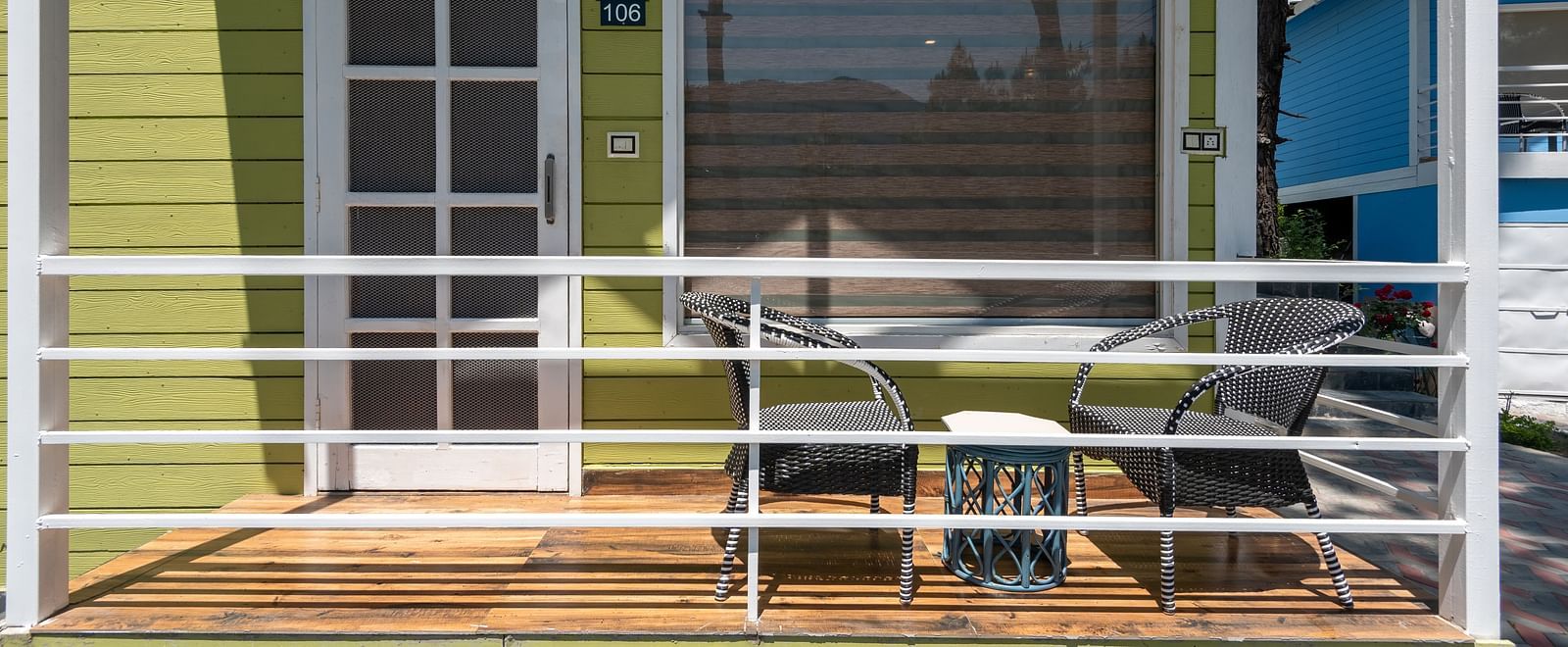  Front porch of a green wooden cottage with white railing, featuring a small outdoor seating area with a table and chairs, and a door and large window behind - DLS Nature Trinket Resort, Dalhousie.