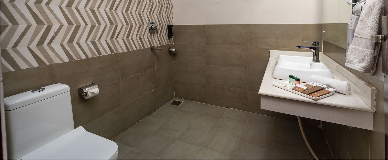Modern bathroom with patterned wall tiles, a toilet, a mounted water heater, a wall-mounted sink with toiletries, and neatly folded towels on a rack - DLS Nature Trinket Resort, Dalhousie.