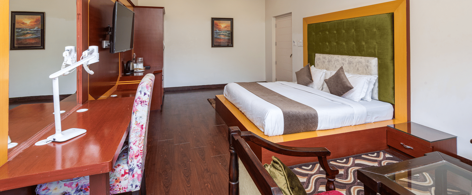 This image displays a spacious hotel room with a large bed, seating area, and desk. Decorated with artwork and includes a ceiling fan | DLS Tehri Club Resort.