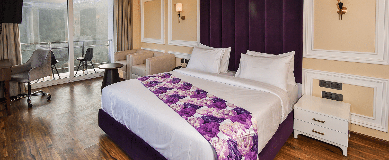 A vibrant bedroom with a large bed featuring purple accents and a bright atmosphere. The room includes large windows that provide scenic views of the hills outside.
