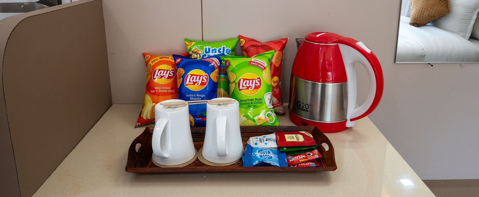 Hotel room refreshment station with a red electric kettle, tea and coffee supplies, two cups, and assorted snack packs arranged neatly on a tray - DLS Nature Trinket Resort, Dalhousie.