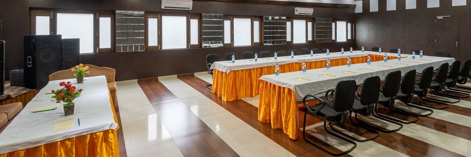 A conference room with a long table, chairs, and decorative flowers. The room has wooden panelling, bright lighting, and windows along the walls - DLS Meghavan Resort, Dharamshala