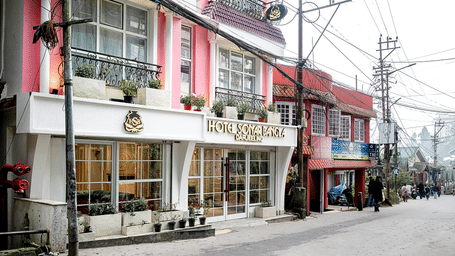 Street view of Hotel Sonar Bangla Darjeeling with a white and red facade, storefront signage, potted plants, and a quiet hill town street.