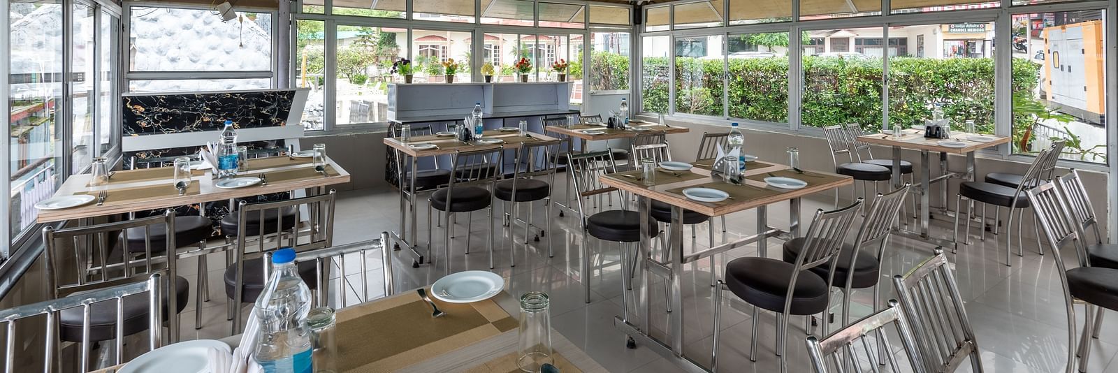 A cafe in Bhagsu with tables and chairs, featuring a green canopy overhead and large windows with views of the outside. The tables are set with plates, glasses, and bottles of water - DLS Meghavan Resort, Dharamshala