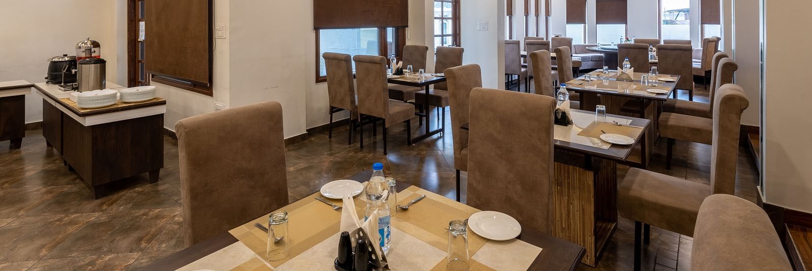 A restaurant with beige seating, tables set with plates, glasses, and cutlery. The space features modern lighting, brown window blinds, and a buffet area - DLS Meghavan Resort, Dharamshala