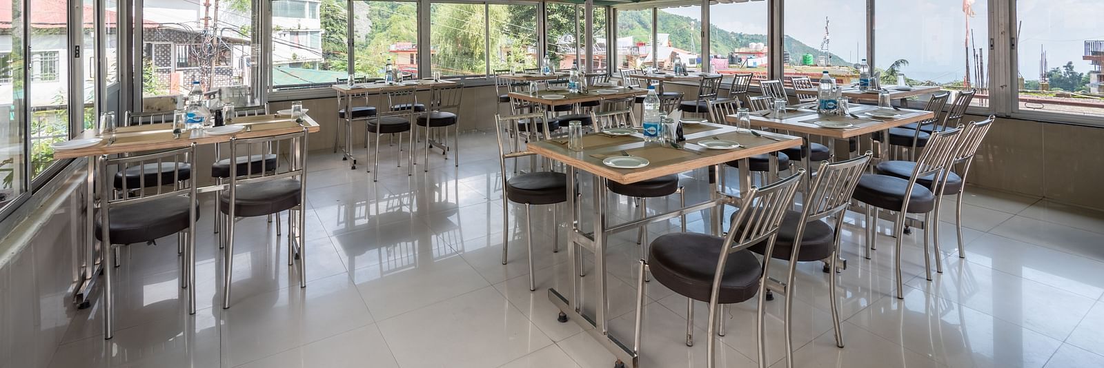 A coffee house with tables and chairs under a green canopy, large windows, and outdoor views. Tables are set with plates and glasses - DLS Meghavan Resort, Dharamshala
