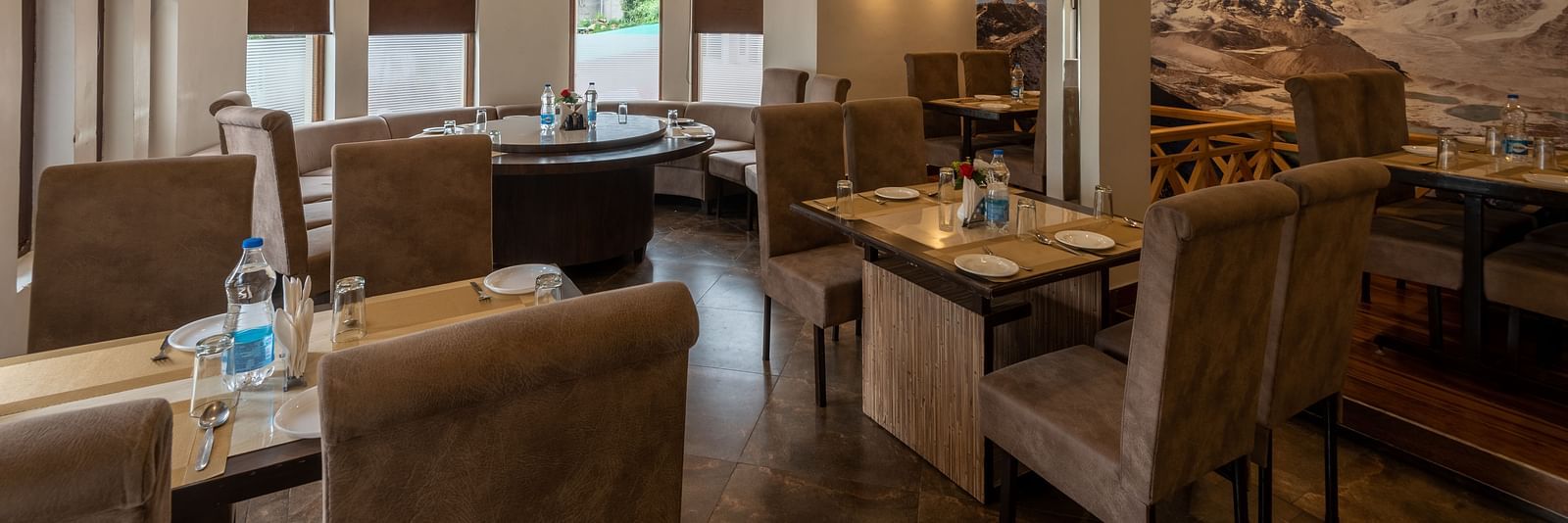 A restaurant with beige seating, tables set with plates, glasses, and cutlery. The space features modern lighting, ceiling fans, and a large mountain mural on the wall -  DLS Meghavan Resort, Dharamshala