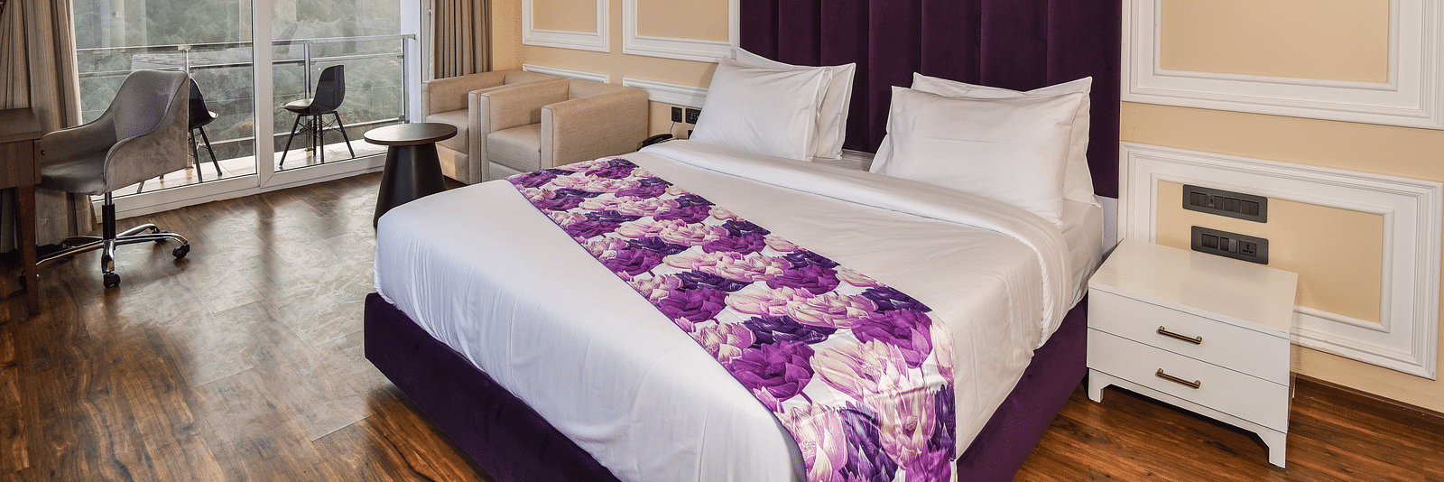 A vibrant bedroom with a large bed featuring purple accents and a bright atmosphere. The room includes large windows that provide scenic views of the hills outside.