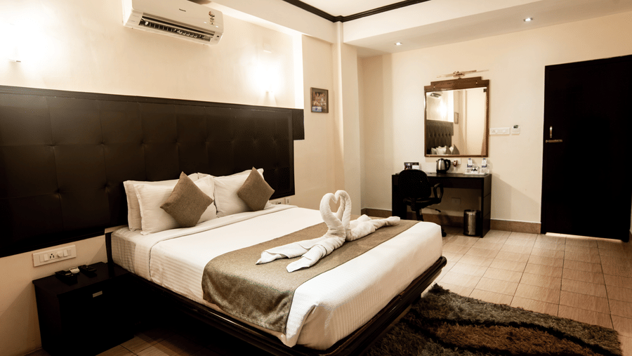 A bedroom at Polo Tura Hotel featuring a neatly made bed with towel swans, a nightstand, an A/C unit, a vanity and a rug.