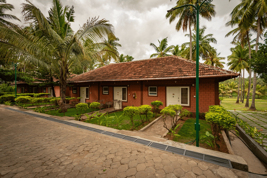 Facade of the River View Suite with gardens, a pathway, and coconut trees under a cloudy sky at Ibex River Resort, Pollachi, one of the best hotels near Aliyar Dam.