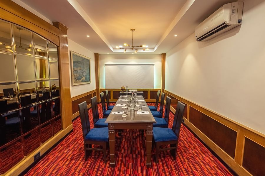 A private dining room with a long wooden table set for a meal, blue upholstered chairs, red patterned carpet, wood paneling, mirrored wall, and a modern chandelier.