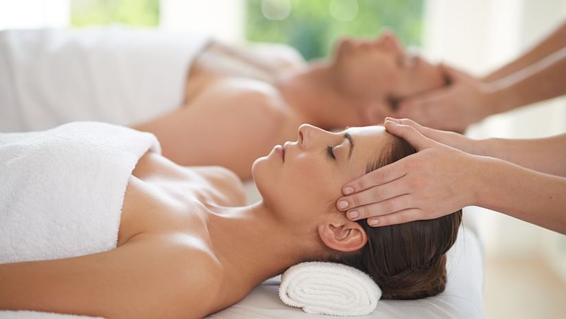 A couple laying on two white massage beds, wearing white towels and facing upwards, while their face is being massaged by two practitioners.