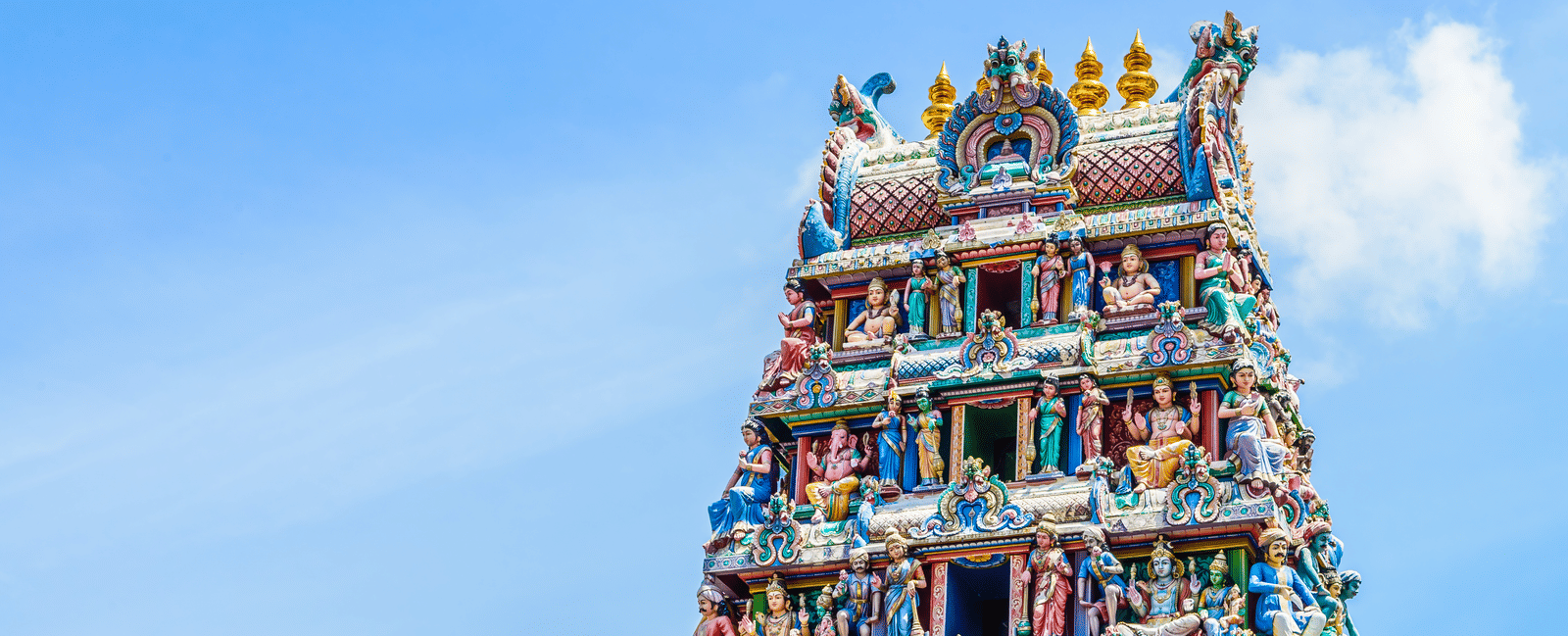 A gopuram stands tall against a bright sky with clouds featuring details of carved figures 