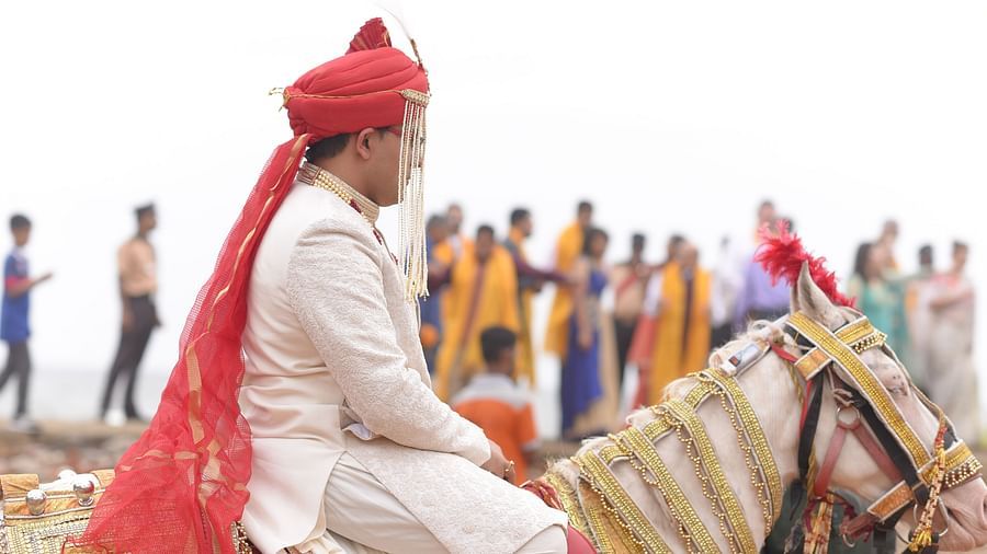 An image of a groom dressed in traditional wedding attire, sitting on a decorated horse, with guests gathered in the background