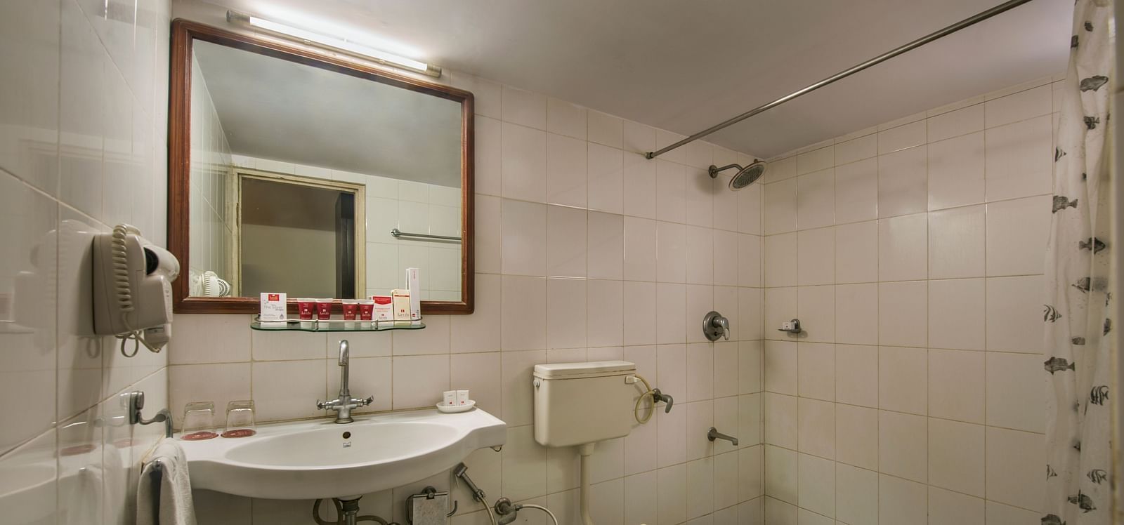 A small bathroom with a white sink, toilet, a mirror with a wooden frame, and a shower area with a patterned curtain - Lords Eco Inn Dahej