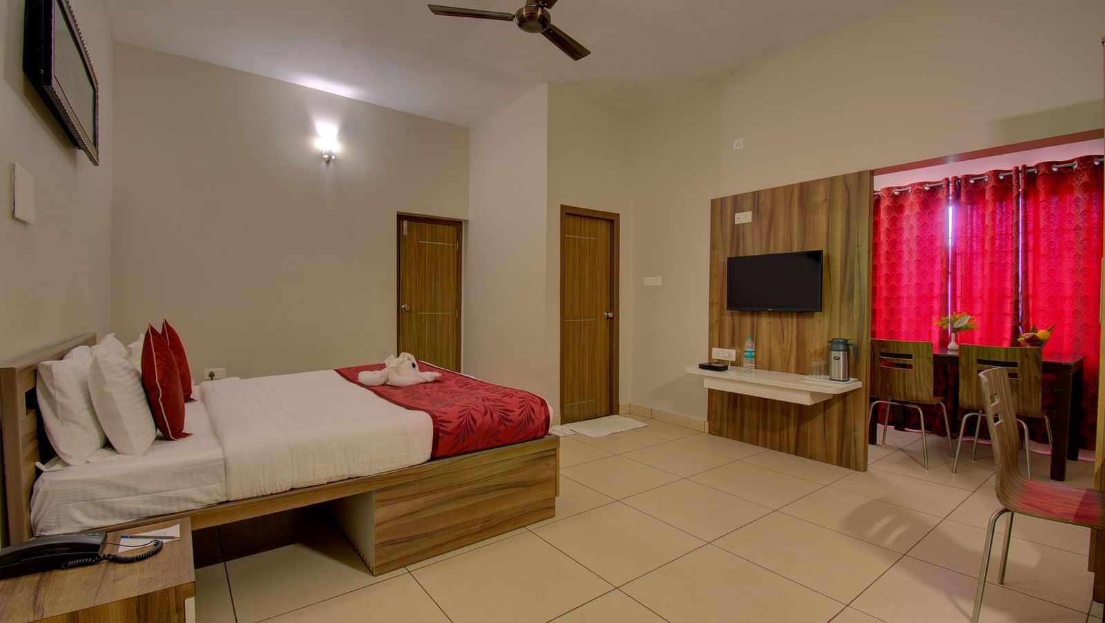 Bedroom with a  huge bed, a side table, chair, table, TV, wooden door and curtained windows | Superior Room | Woodstock Resorts, Coorg