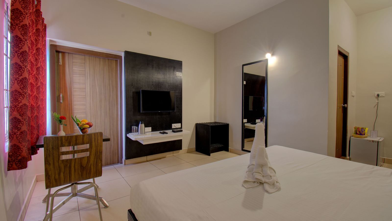A bedroom with a bed, chair, TV unit, curtained windows, mirror and wooden door | Superior Room | woodstock resorts, Coorg