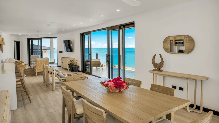 A dining area with a dining table with a flower bowl and chairs, two lounge chairs, a recliner, a side table, and a glass sliding door leading to a patio with a sea view at the Tamarind Hills Resort and Villas, Antigua.
